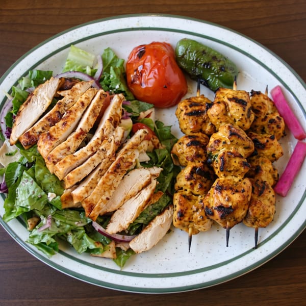 Grilled chicken breast slices, chicken skewers, roasted tomato, green pepper, and pickled turnips on a plate.
